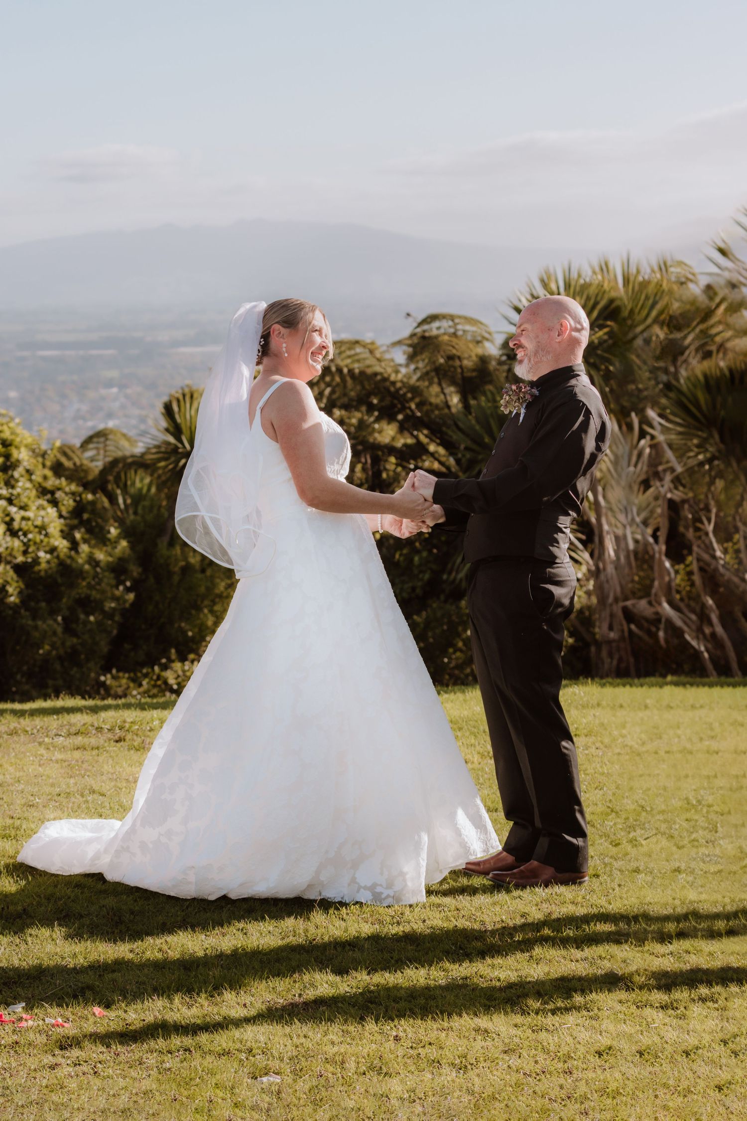 Couple in wedding attire dance together in a sequence of joyful movements on a grassy hilltop with mountains in background.