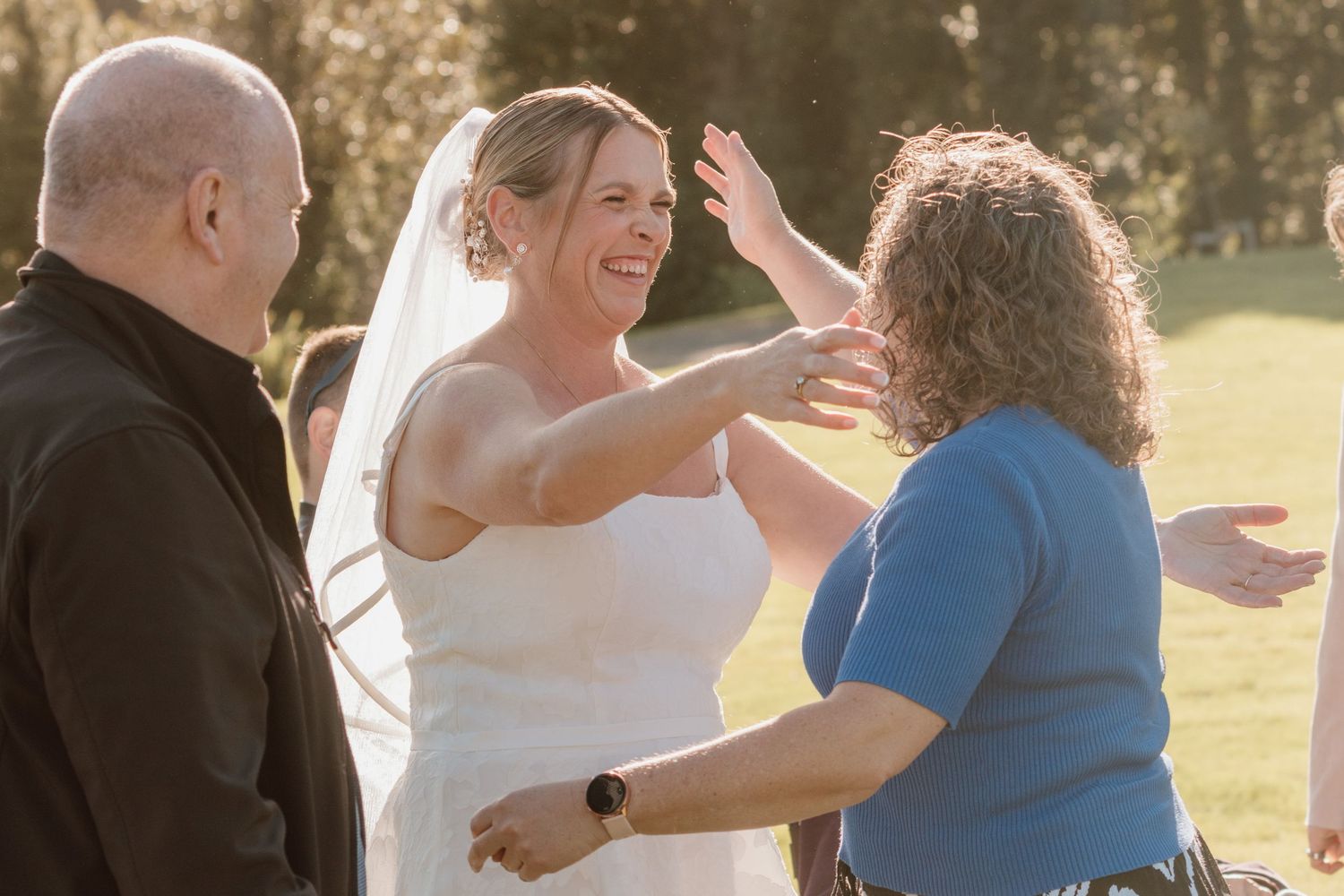 A bride in a white wedding dress shares a joyful moment with two guests during an outdoor ceremony.