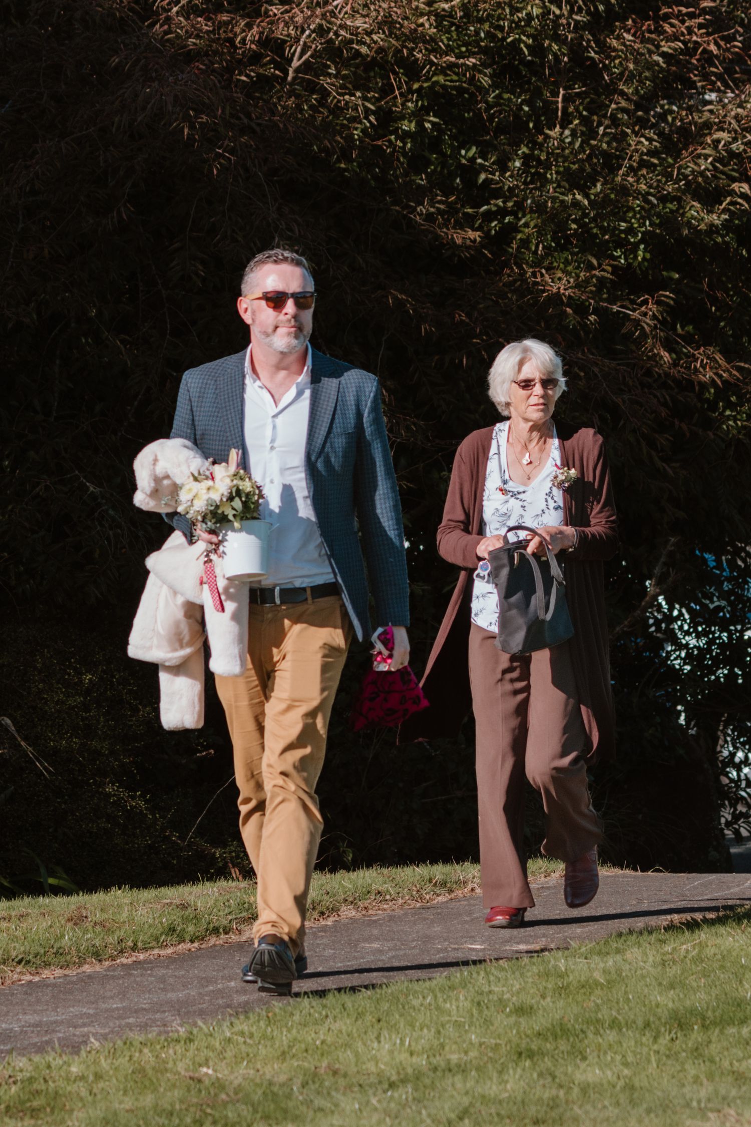 Elegant couple walking outdoors, carrying teddy bears in formal attire on a sunny day against greenery background.