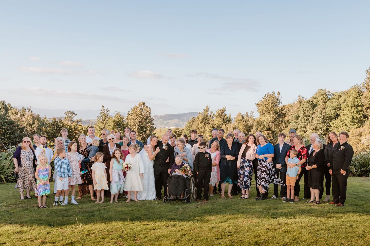 Wedding guests and family members pose together on scenic lawn with mountain vista at sunset.