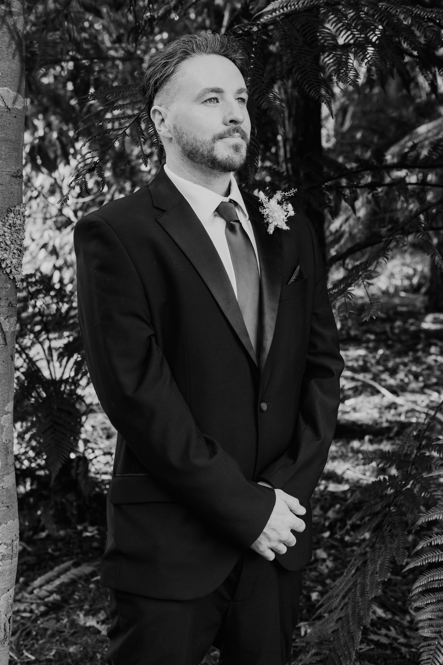 Black and white portrait of a person in a formal suit and tie standing against a leafy garden background.
