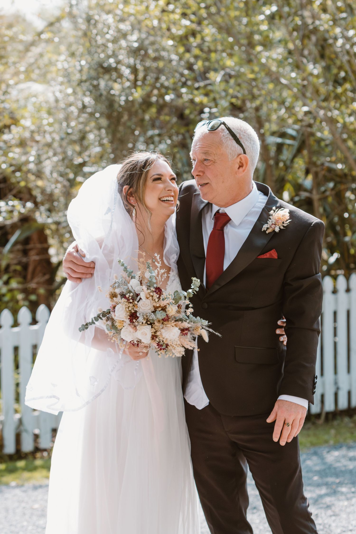 A bride in a white dress and veil shares a sweet moment outdoors with a man in a dark suit and red tie.