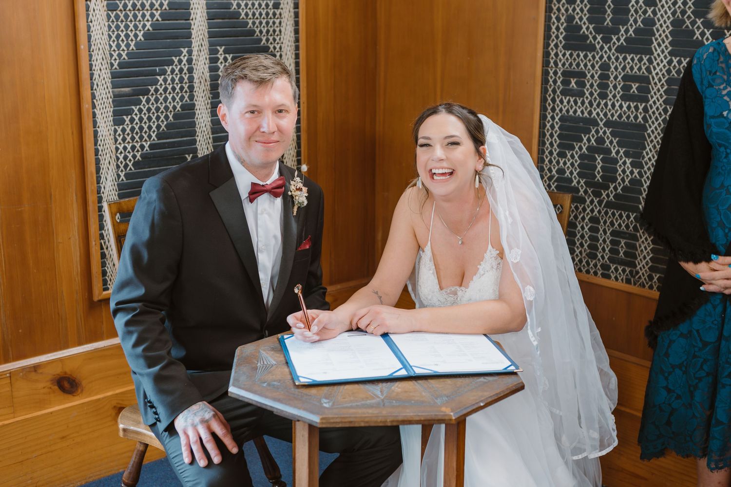Wedding couple laughs with joy while signing their marriage certificate at wooden table during ceremony.