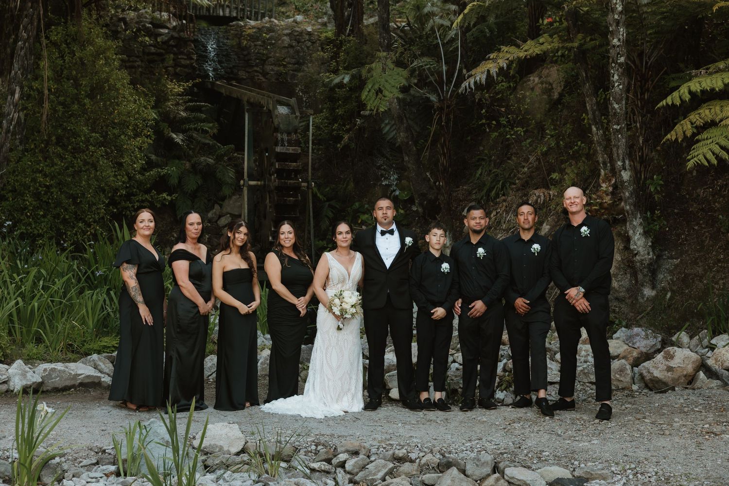 A wedding party in black and white attire poses outdoors against a natural forest backdrop with rocks and greenery.