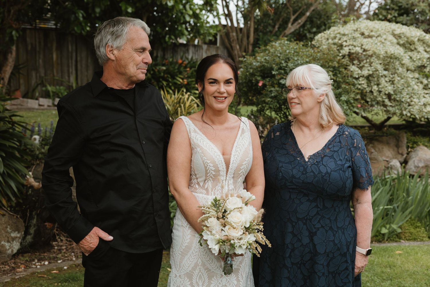 A bride in a white lace wedding dress poses with family members in a garden setting.