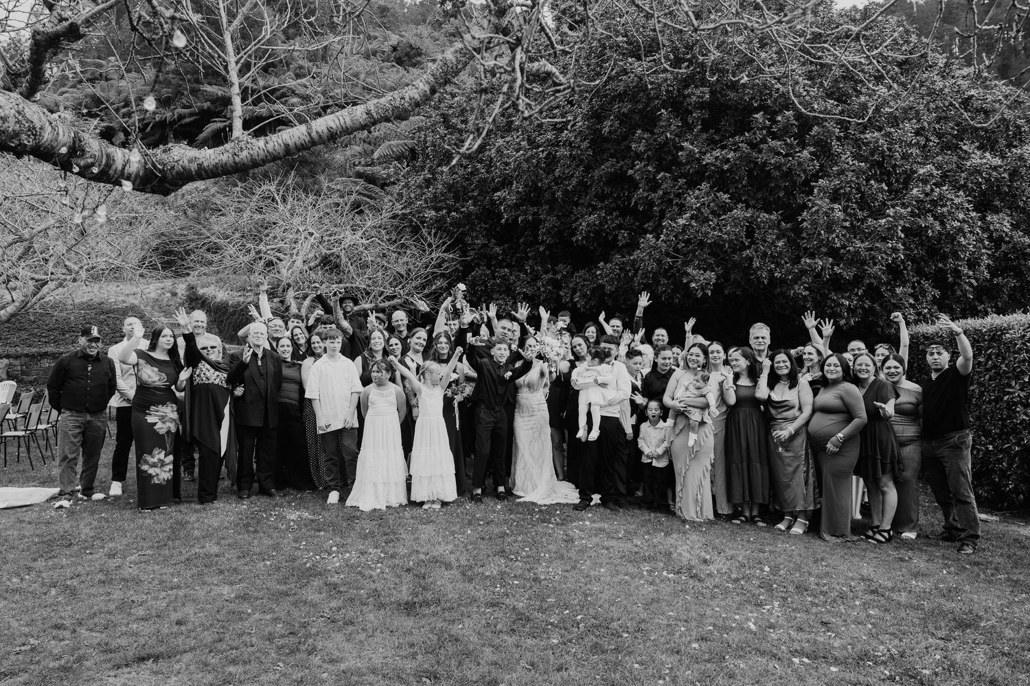 Black and white photo of a large wedding party gathered outdoors on a lawn with trees in the background.