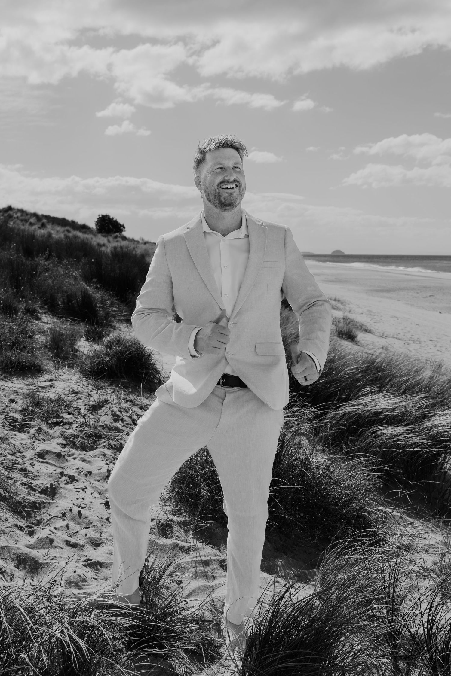 Black and white photo of a smiling man in a suit standing on sandy beach dunes with ocean in background.