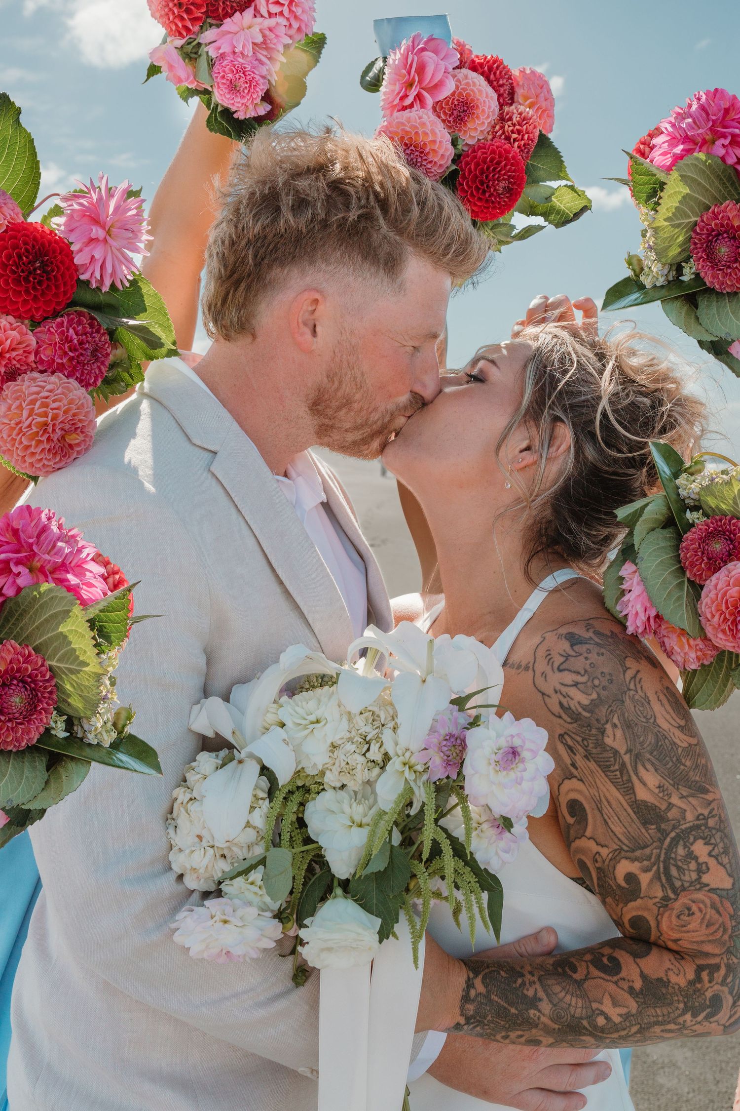Bride and groom share a kiss under a floral arch with pink dahlias, bride holding white bouquet and showing tattoo sleeve.