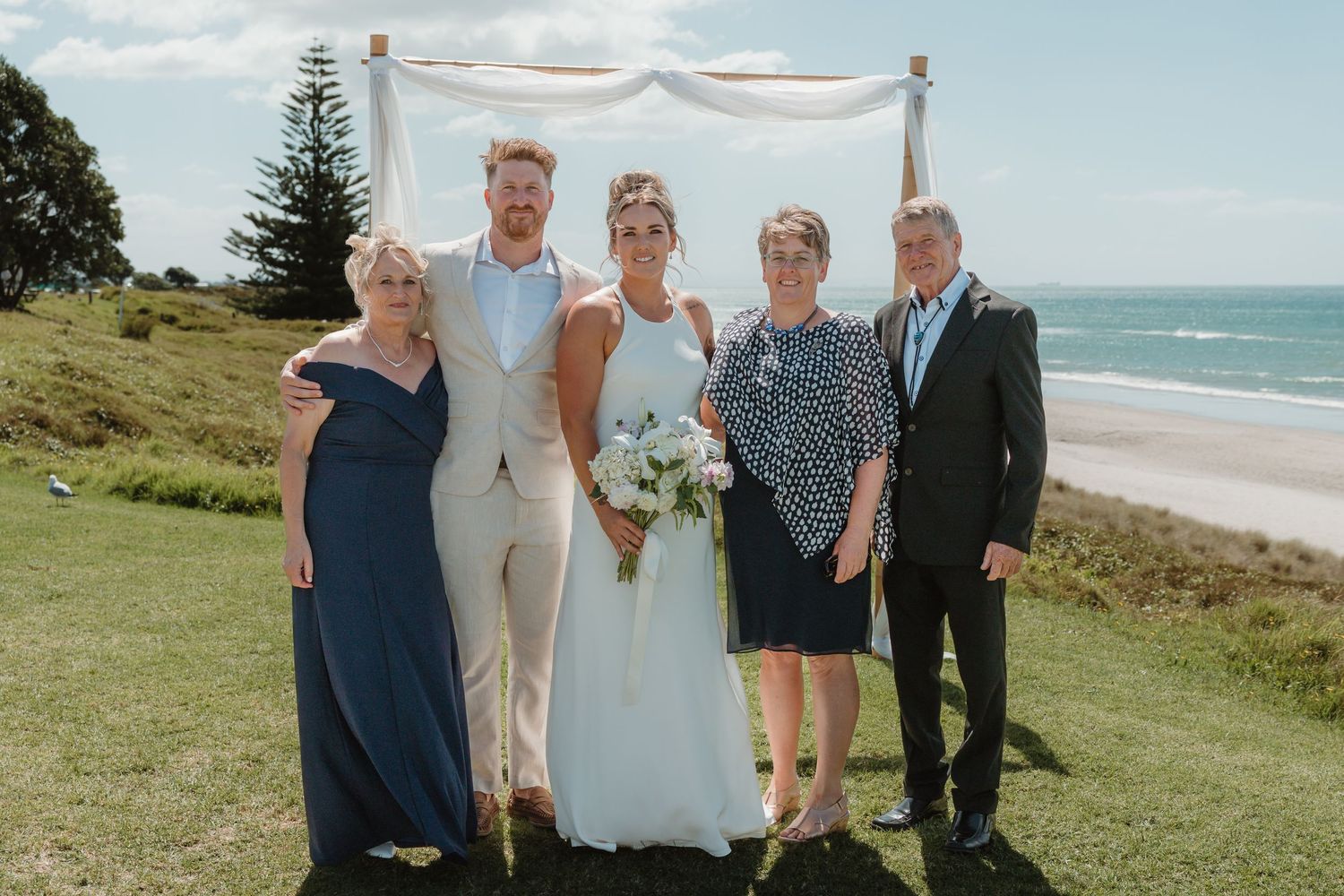 Wedding party of five posing outdoors by the ocean under a draped arch on a sunny day.