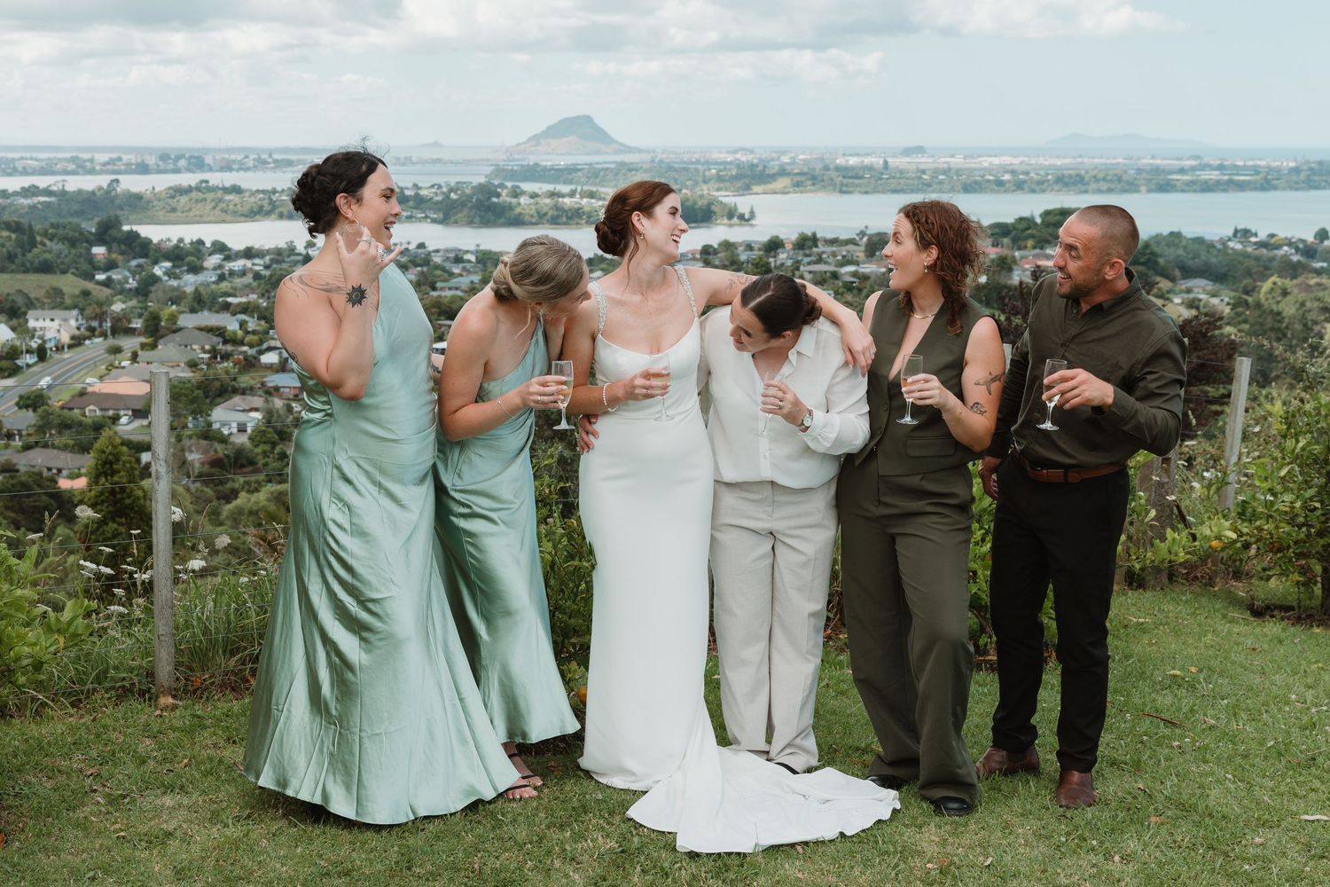 Bride in white gown with bridesmaids in sage green dresses celebrating outdoors with scenic coastal town views.