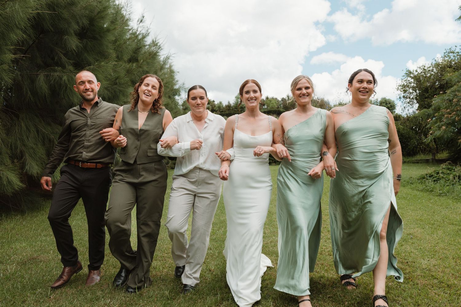 Bride in white gown walks with bridesmaids in sage green dresses and groomsman outdoors on lush green lawn.