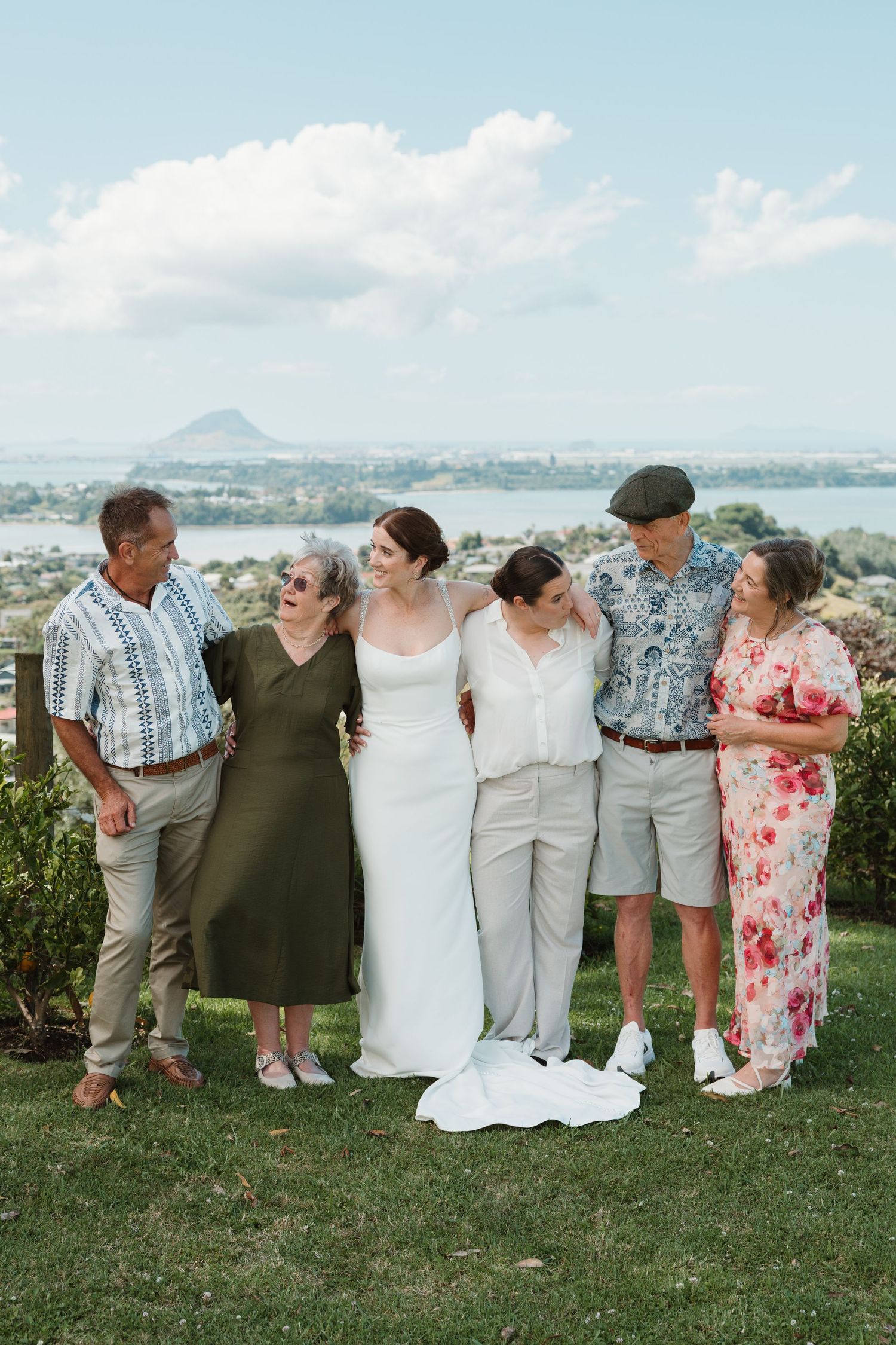 Bride in white gown poses with family outdoors at scenic waterfront wedding venue under blue sky.
