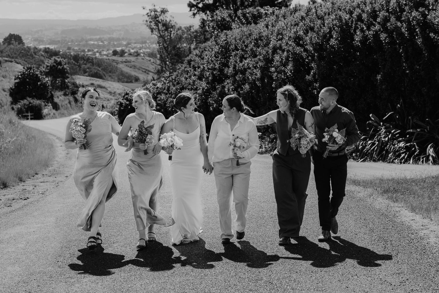 Black and white photo of a bridal party walking and laughing together on a scenic road surrounded by trees.