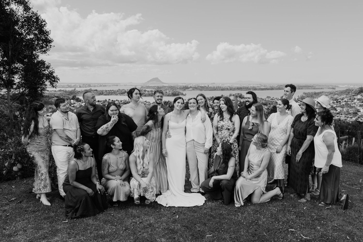 Black and white wedding photo of bride and groom with large bridal party outdoors, scenic landscape in background.