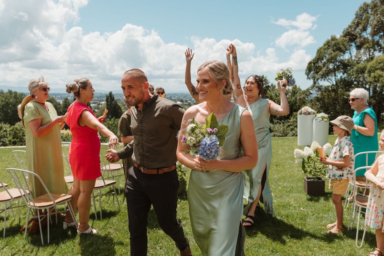 Bride in mint green dress walks with groom outdoors at sunny garden wedding ceremony with guests celebrating.