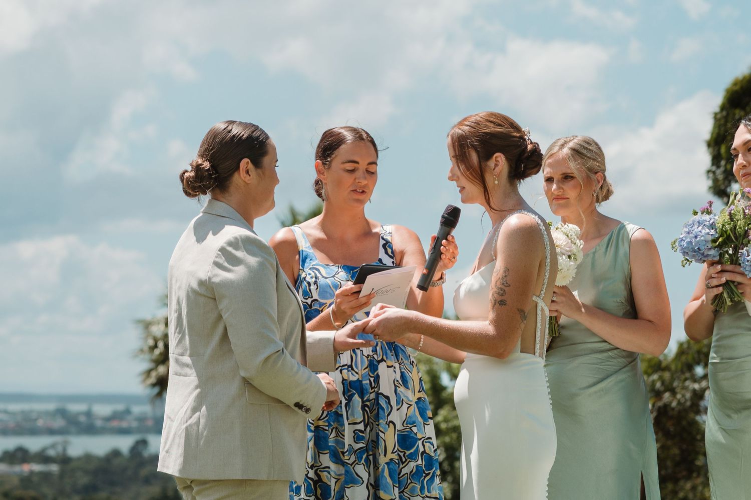 Two brides exchange vows outdoors at a scenic wedding ceremony, officiated by a woman in a floral dress.