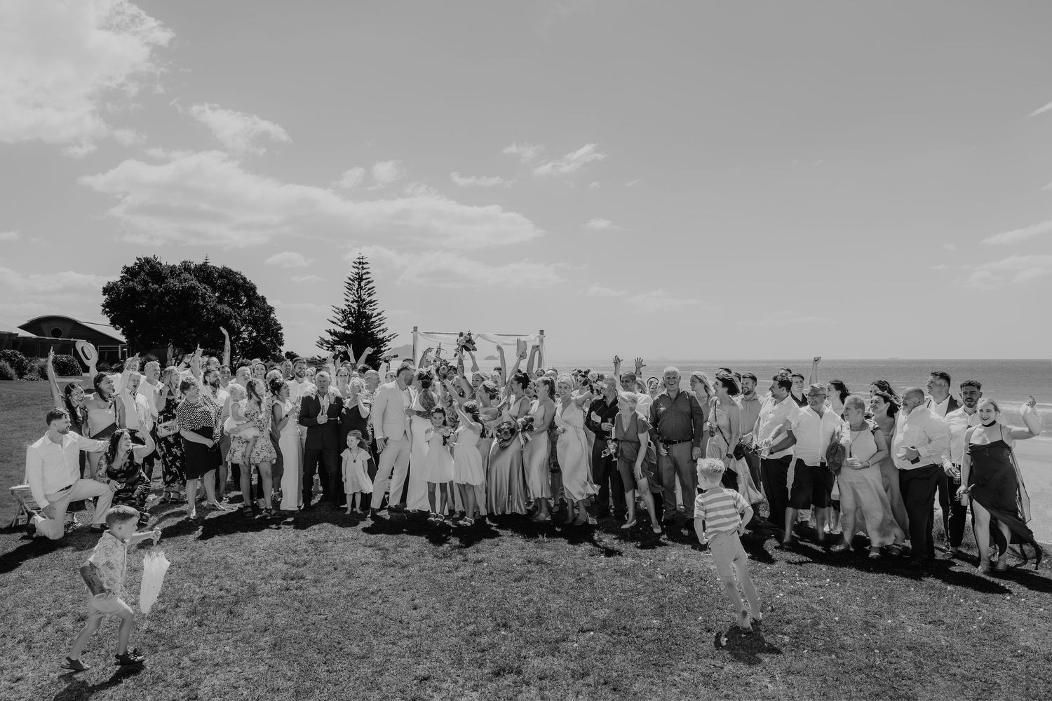 Black and white photo of a large wedding group celebrating outdoors on a grassy field near the ocean.