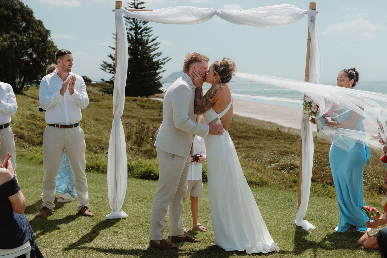 Bride and groom share first kiss at outdoor coastal wedding ceremony under white draped arch.