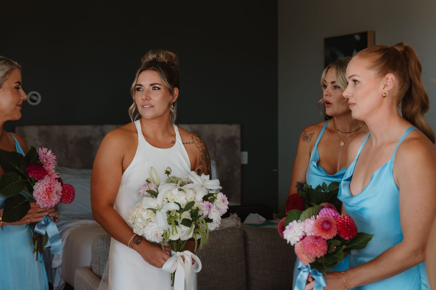 Bride in white dress holds bouquet with bridesmaids in blue dresses during wedding preparations indoors.