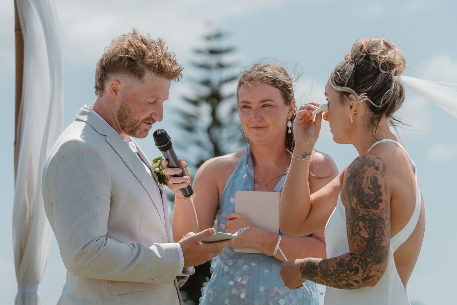 Emotional outdoor wedding ceremony with groom reading vows as bride wipes tears, officiant holding papers nearby.