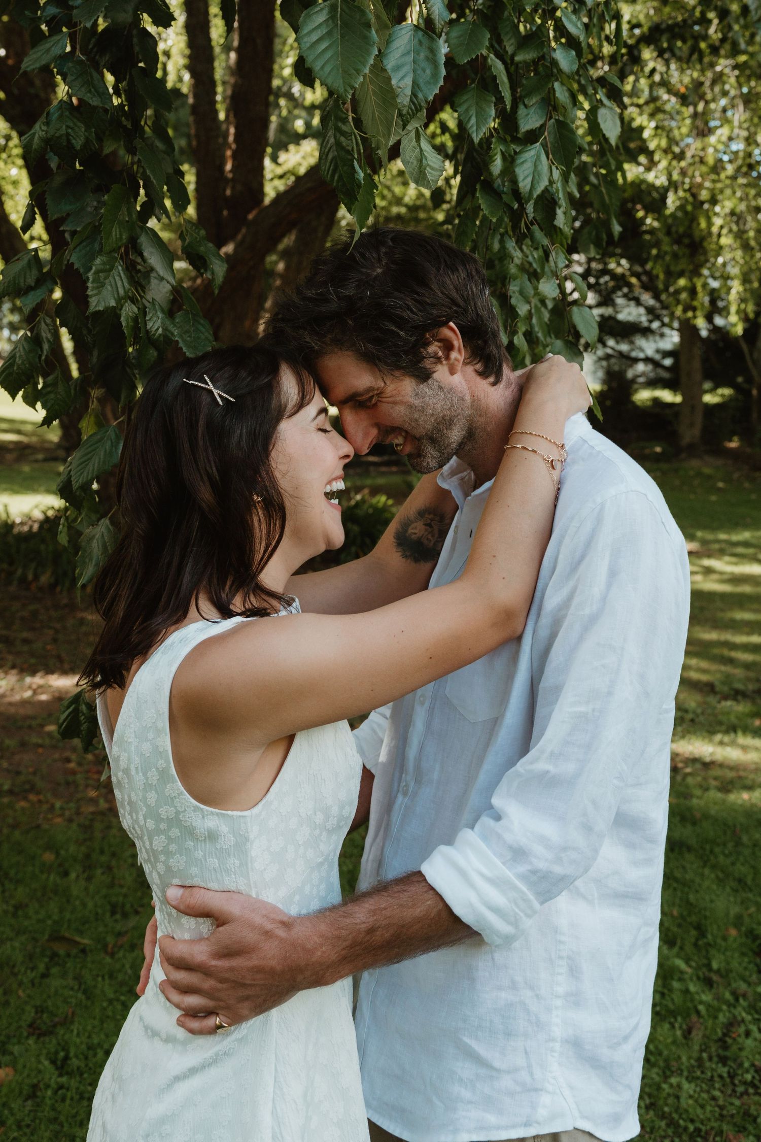 A couple embraces and smiles at each other in a lush green park, wearing white outfits in a romantic outdoor setting.