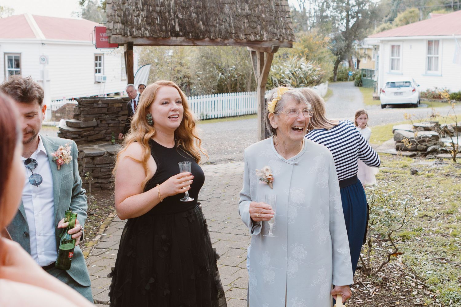 Wedding reception guests gather outdoors near a rustic white building.