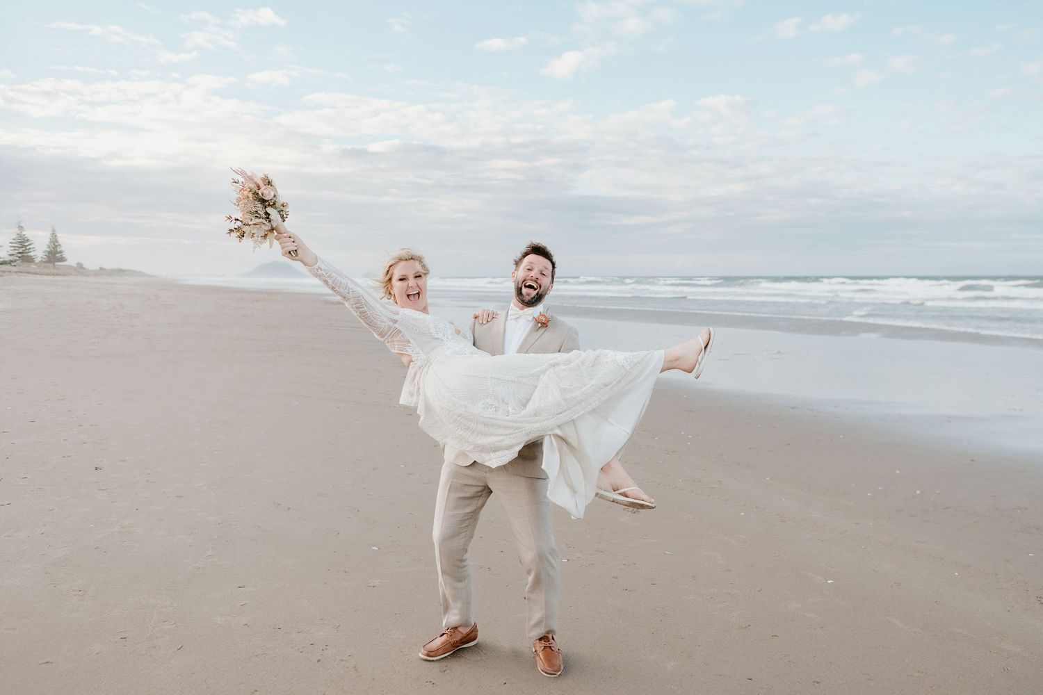 Couple dancing playfully on sandy beach in wedding attire. Papamoa Surf Lifesaving Club Wedding