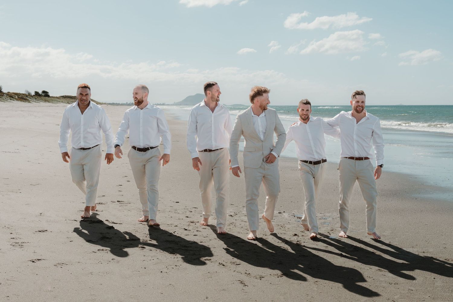 Six groomsmen in white shirts and beige pants walking together on a sandy beach on a sunny day.