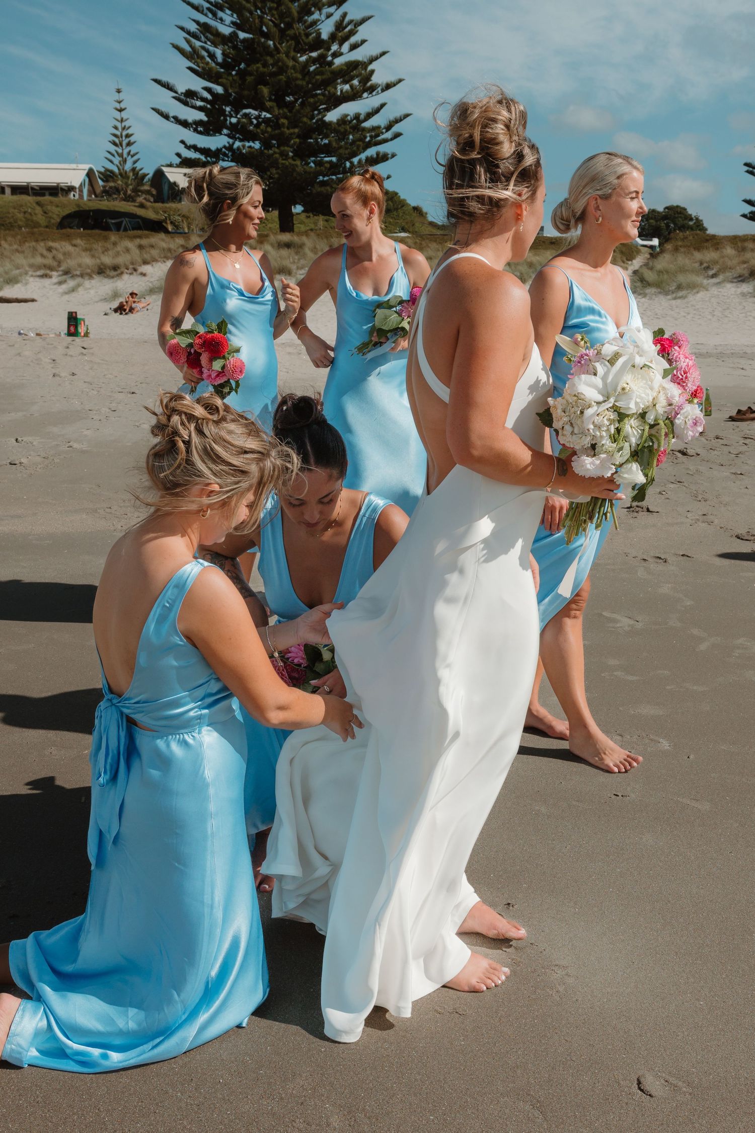 Bride in white gown with bridesmaids in blue dresses on a sandy beach, holding pink and white bouquets.