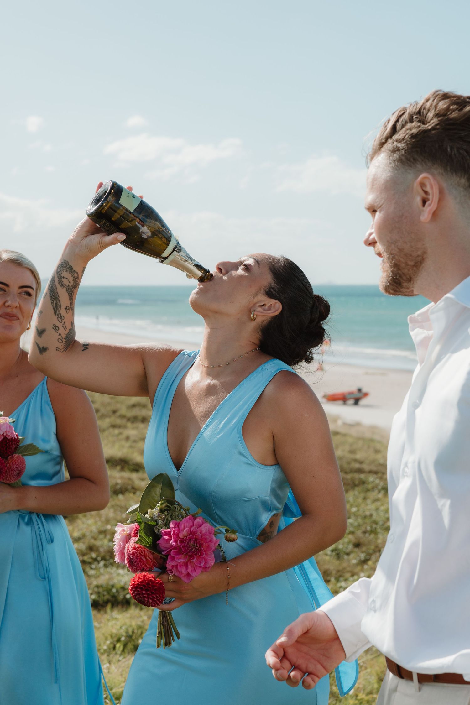 Bridesmaid in blue dress drinks champagne bottle at beach wedding ceremony with bridal party.