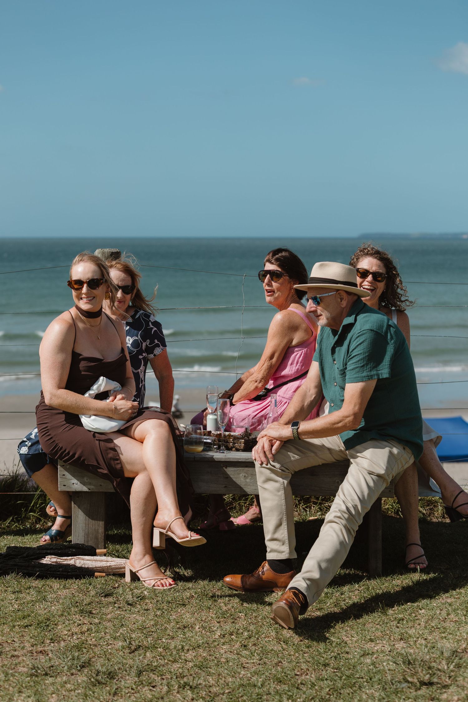 Group of five friends enjoying a sunny beach picnic at a seaside table, wearing sunglasses on a bright clear day.