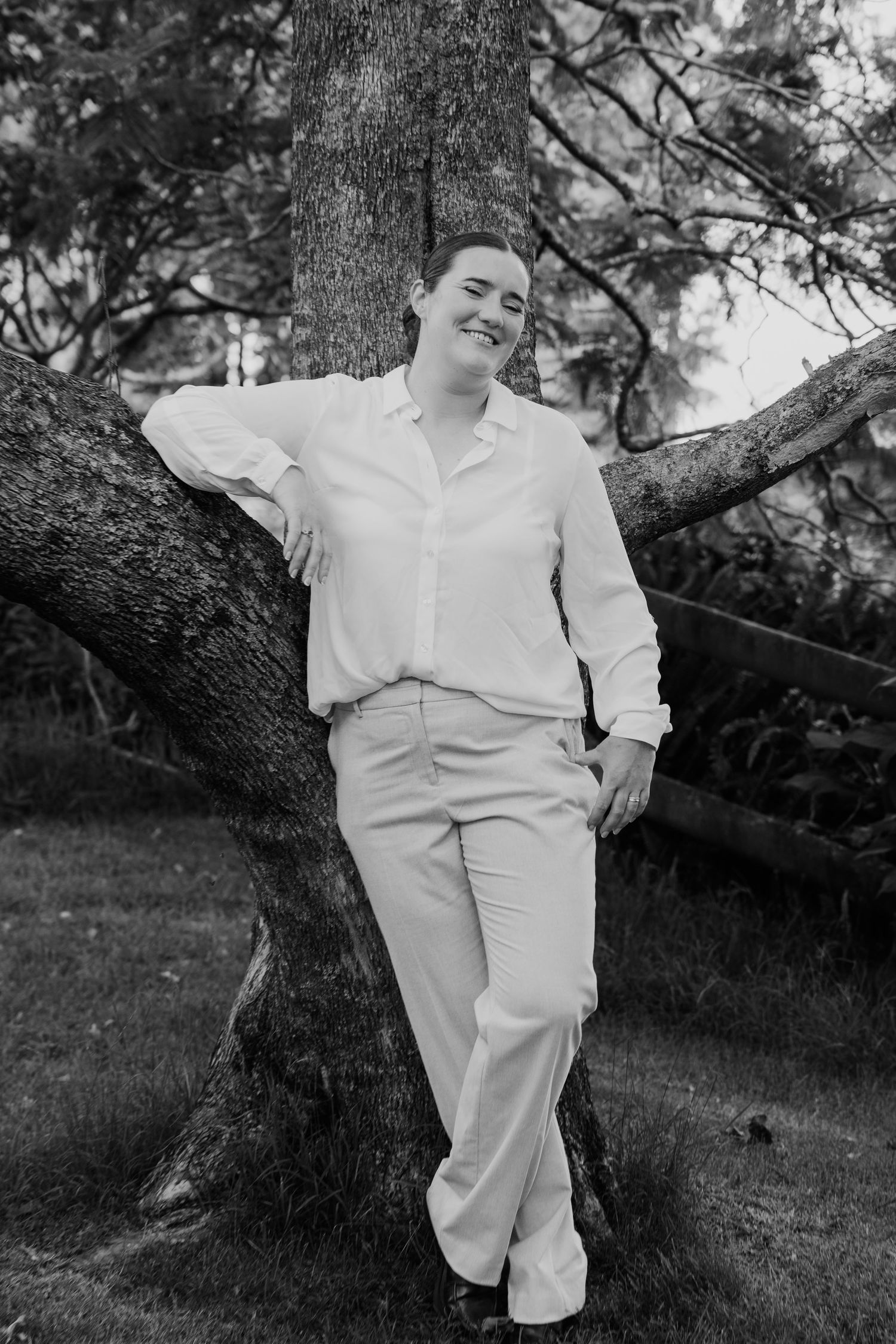 Black and white portrait of a smiling person leaning against a large tree in a lush garden, wearing a white shirt and pants.