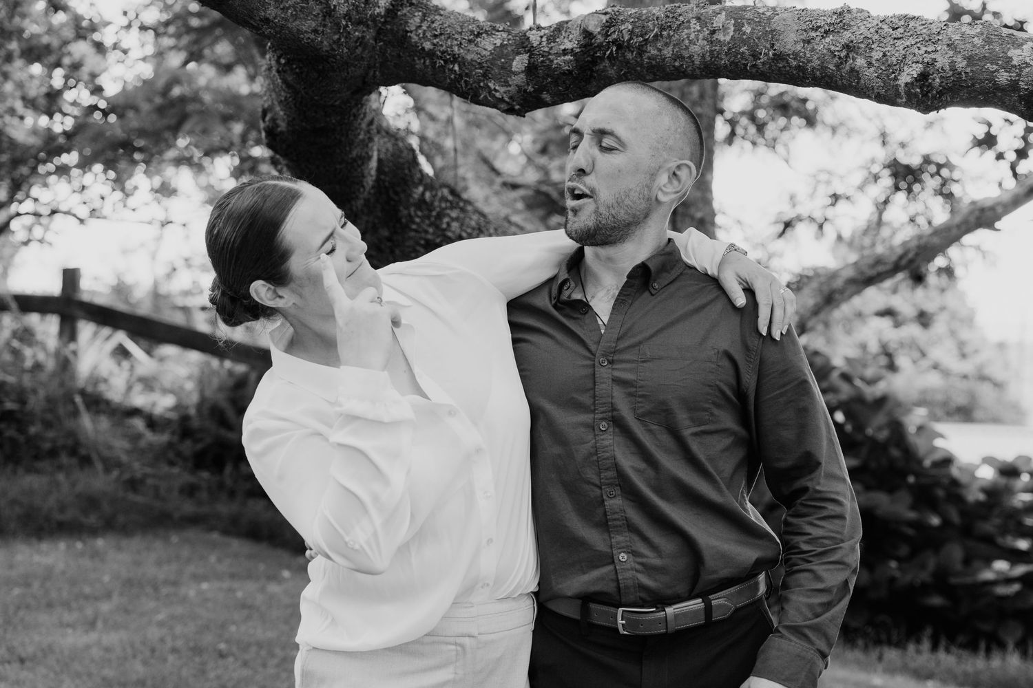Black and white photo of a couple laughing together outdoors under a large tree during an engagement session.