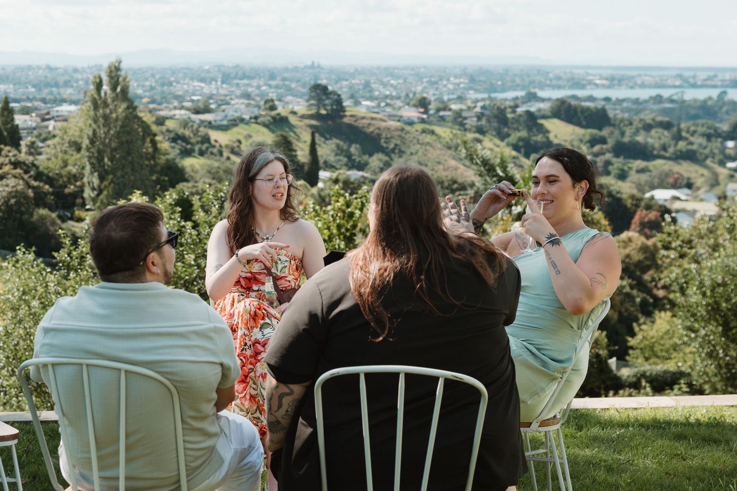 Four friends enjoying drinks outdoors on a sunny hilltop with a scenic city view in the background.