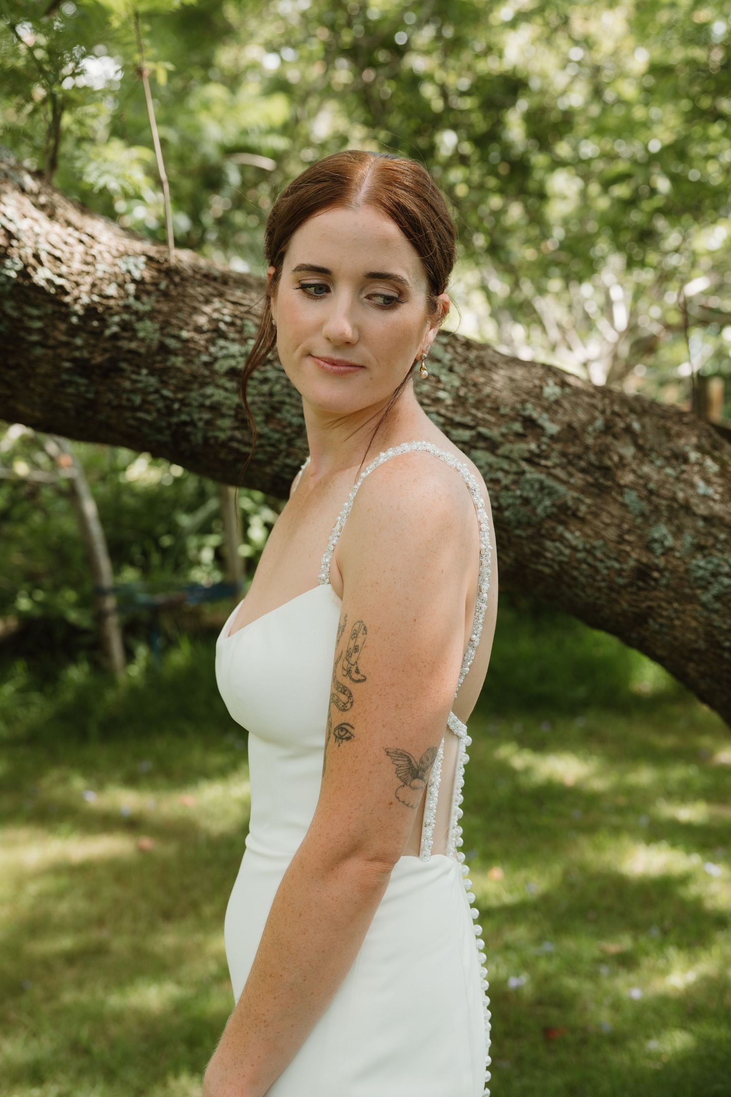 Bride in white gown with tattoos poses outdoors near a large tree trunk in a lush green garden setting.