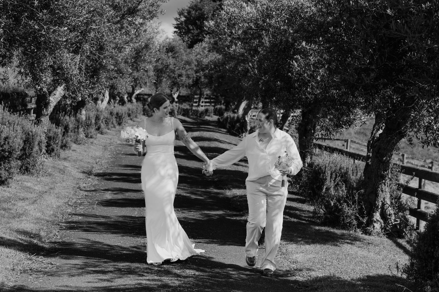 Black and white photo of a bride and groom holding hands, walking joyfully down a tree-lined path on their wedding day.