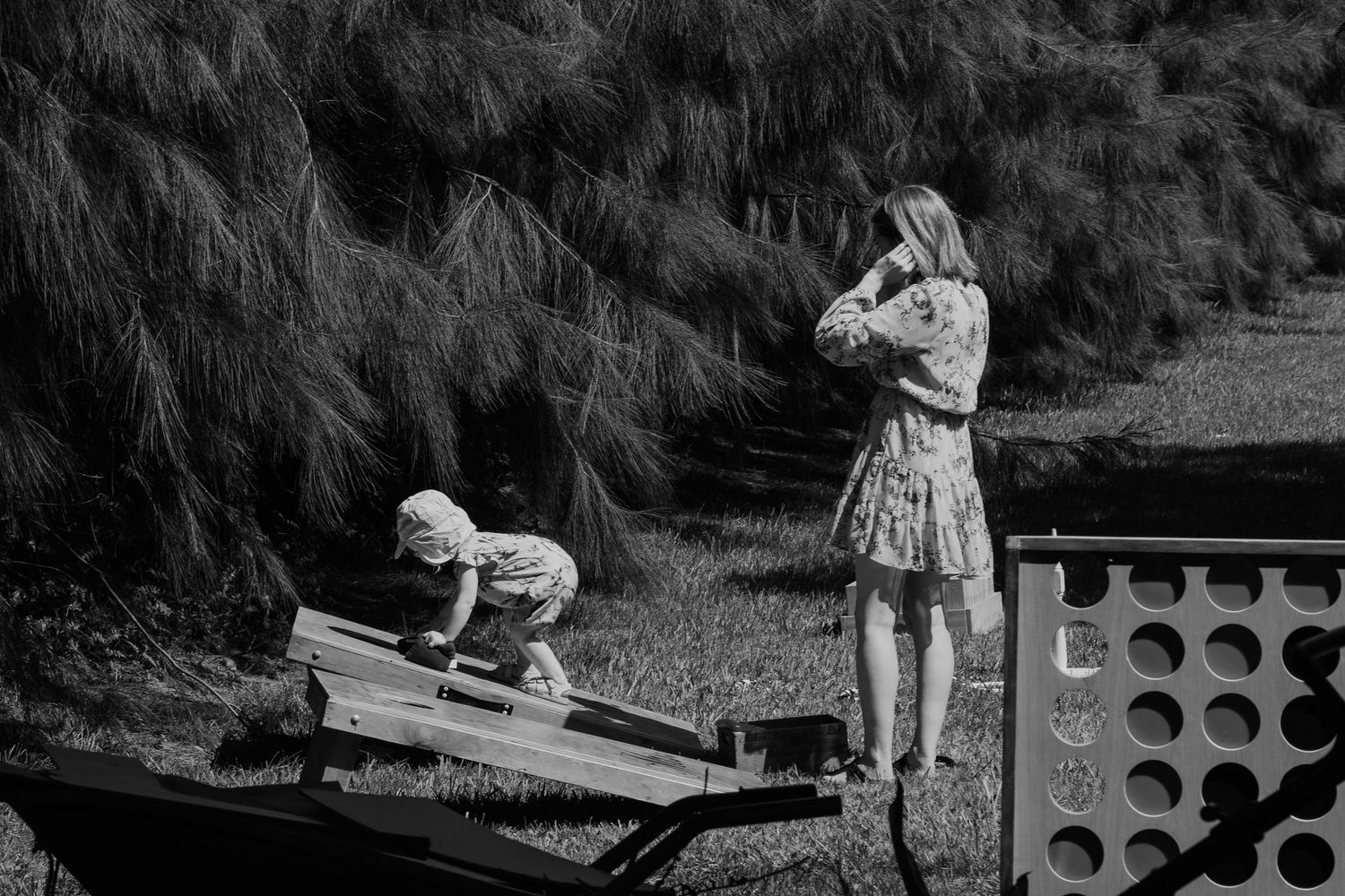 Woman in floral dress watches child play on seesaw at outdoor playground surrounded by tall grass.