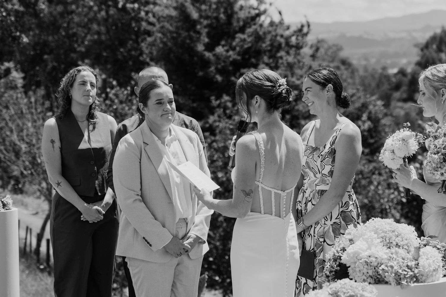 Black and white outdoor wedding ceremony with bride in lace gown, partner in suit, and bridesmaids holding bouquets.