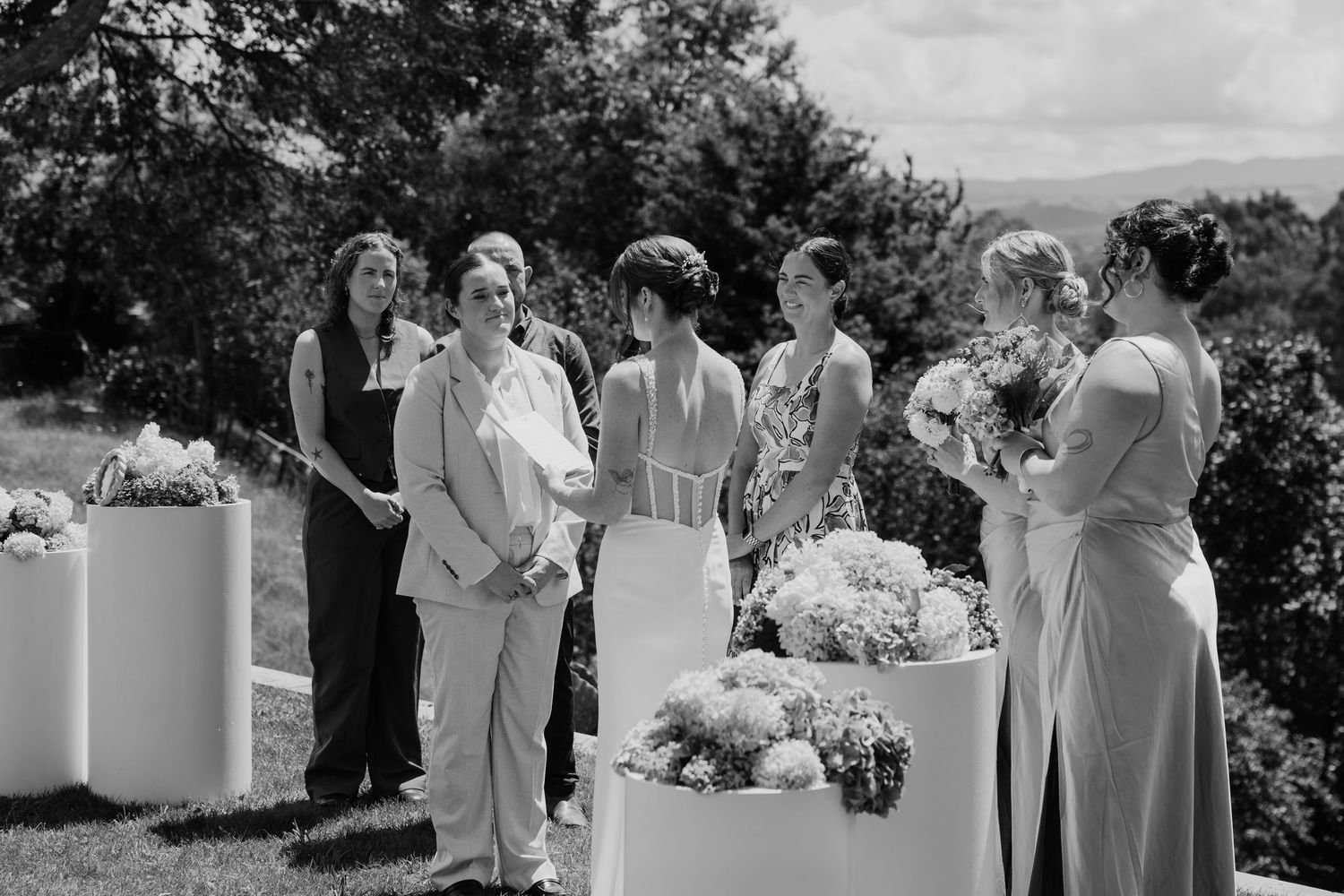 Black and white photo of bridesmaids in elegant gowns gathered outdoors at a wedding ceremony with floral arrangements.