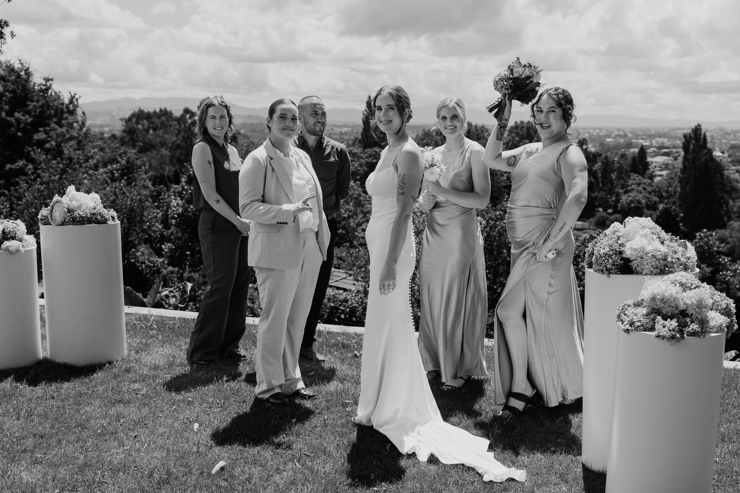 Black and white photo of a bride with bridesmaids in elegant gowns posing outdoors at a scenic wedding venue.