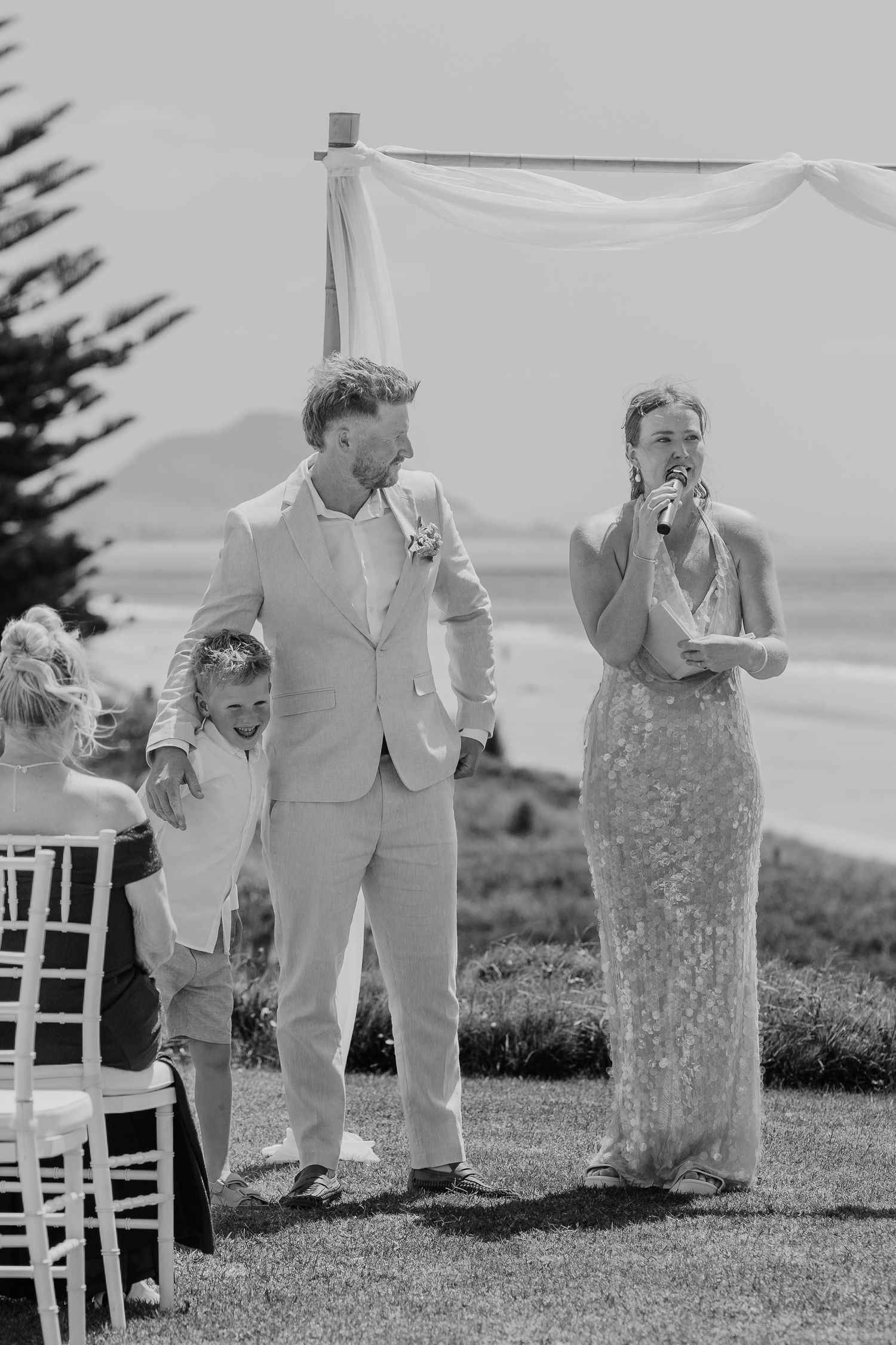 Black and white outdoor wedding ceremony with groom, child, and bride in sequin dress near beach arbor.