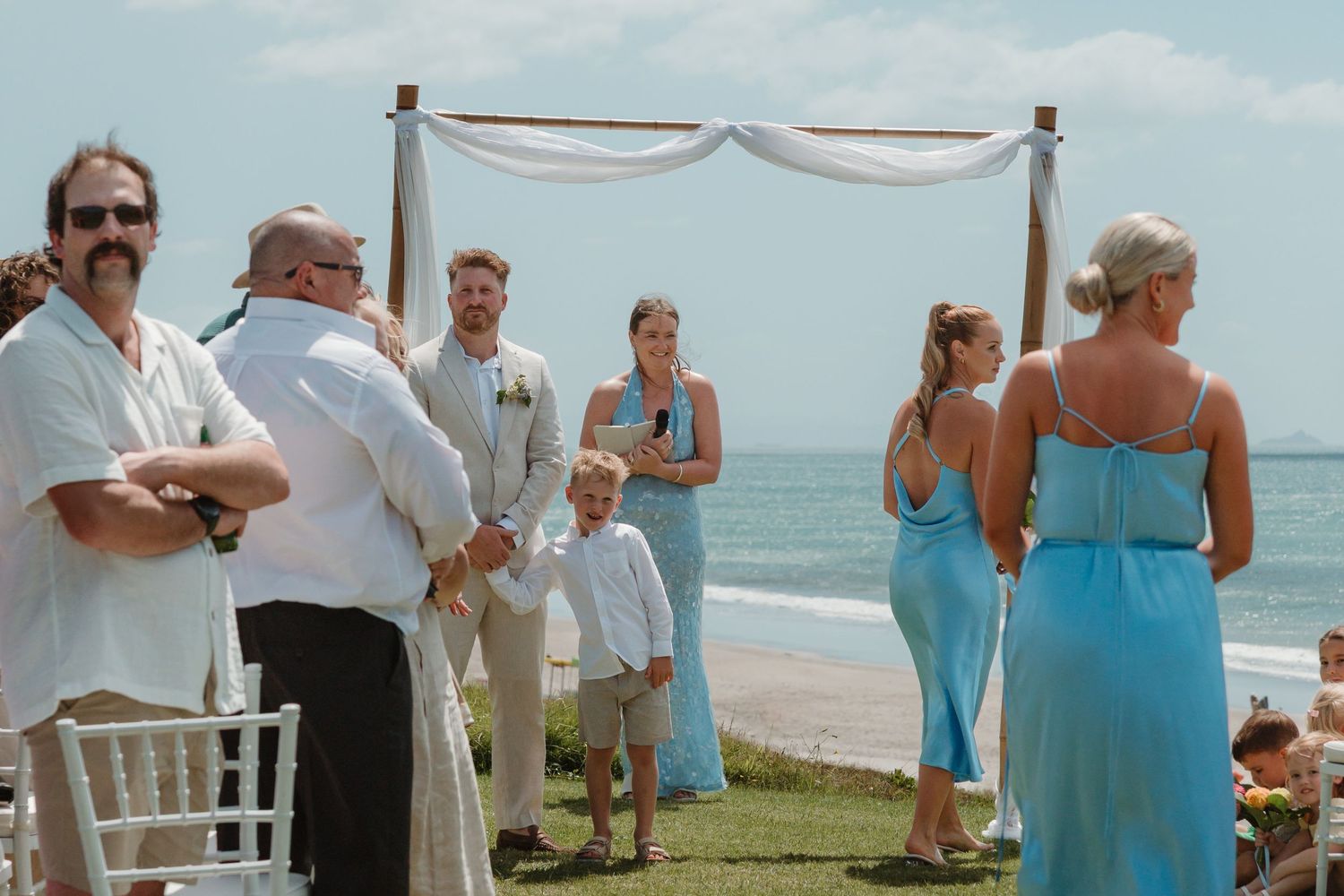 Beach wedding ceremony with couple at bamboo arch, guests in blue attire, ocean backdrop on sunny day.