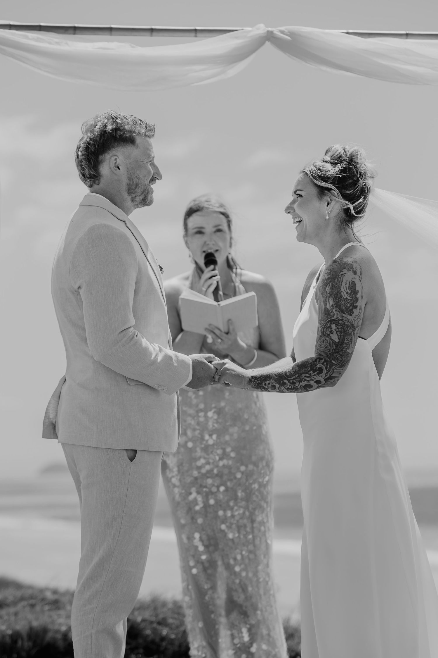 Black and white photo of a couple exchanging vows at an outdoor wedding ceremony with an officiant reading from a book.