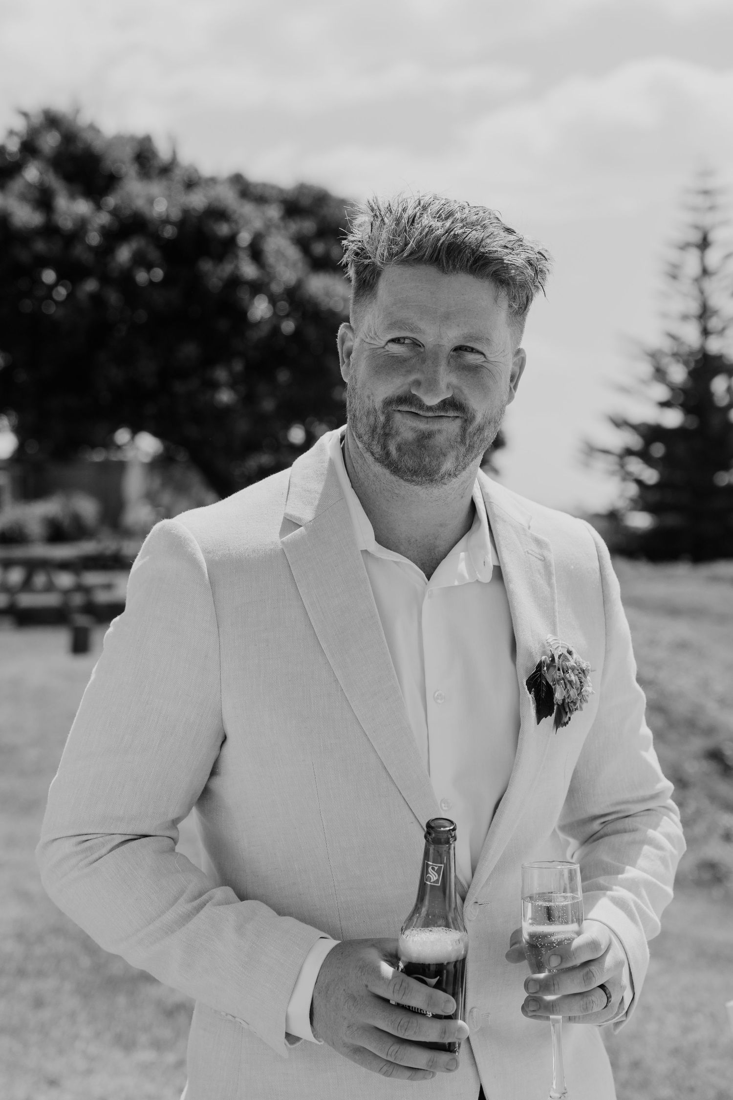 Black and white photo of a smiling bearded groom in a light suit holding a beer bottle and champagne glass outdoors.