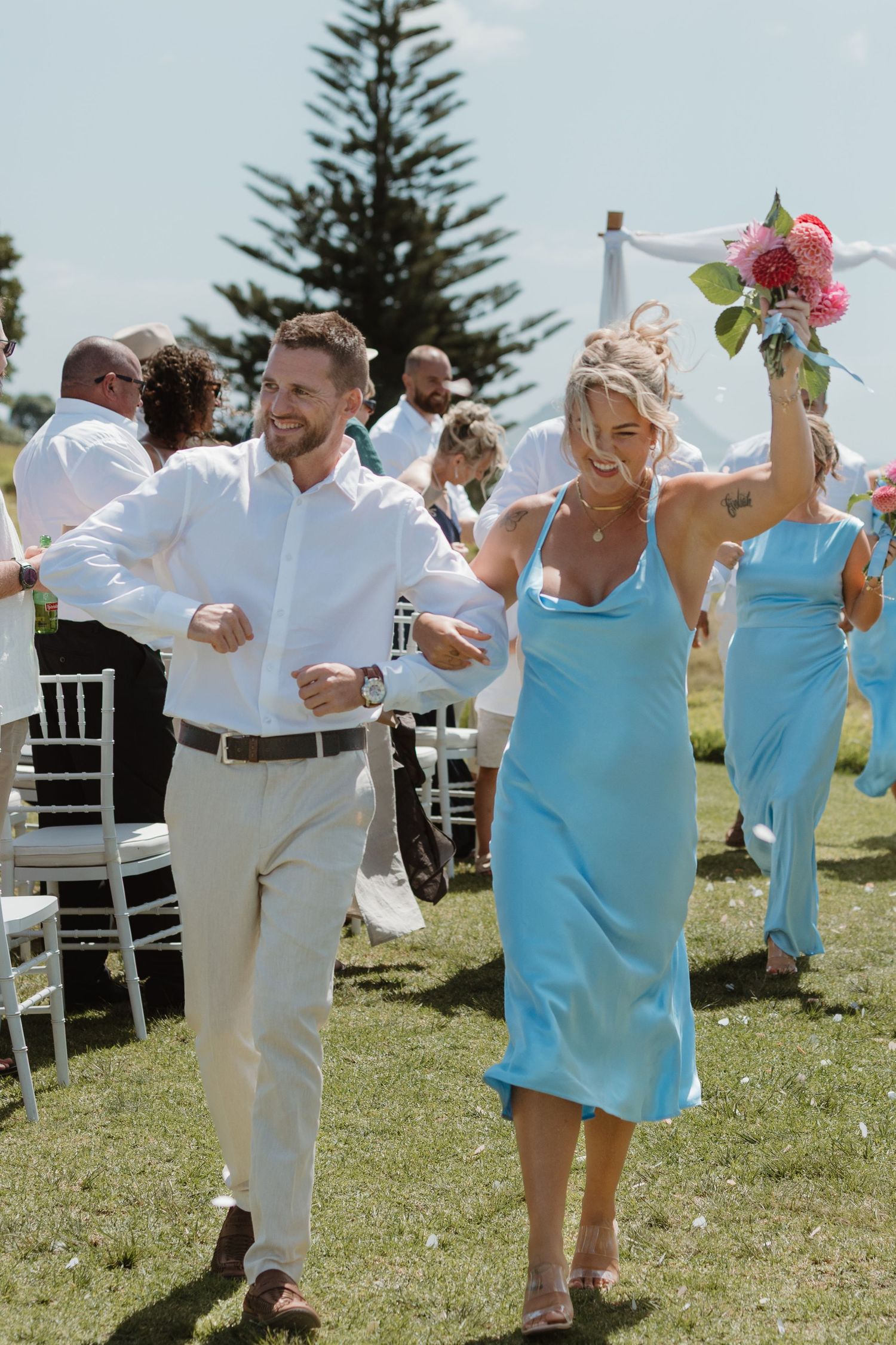 A bridesmaid in a blue dress raises a bouquet while walking with a groomsman at an outdoor wedding ceremony.