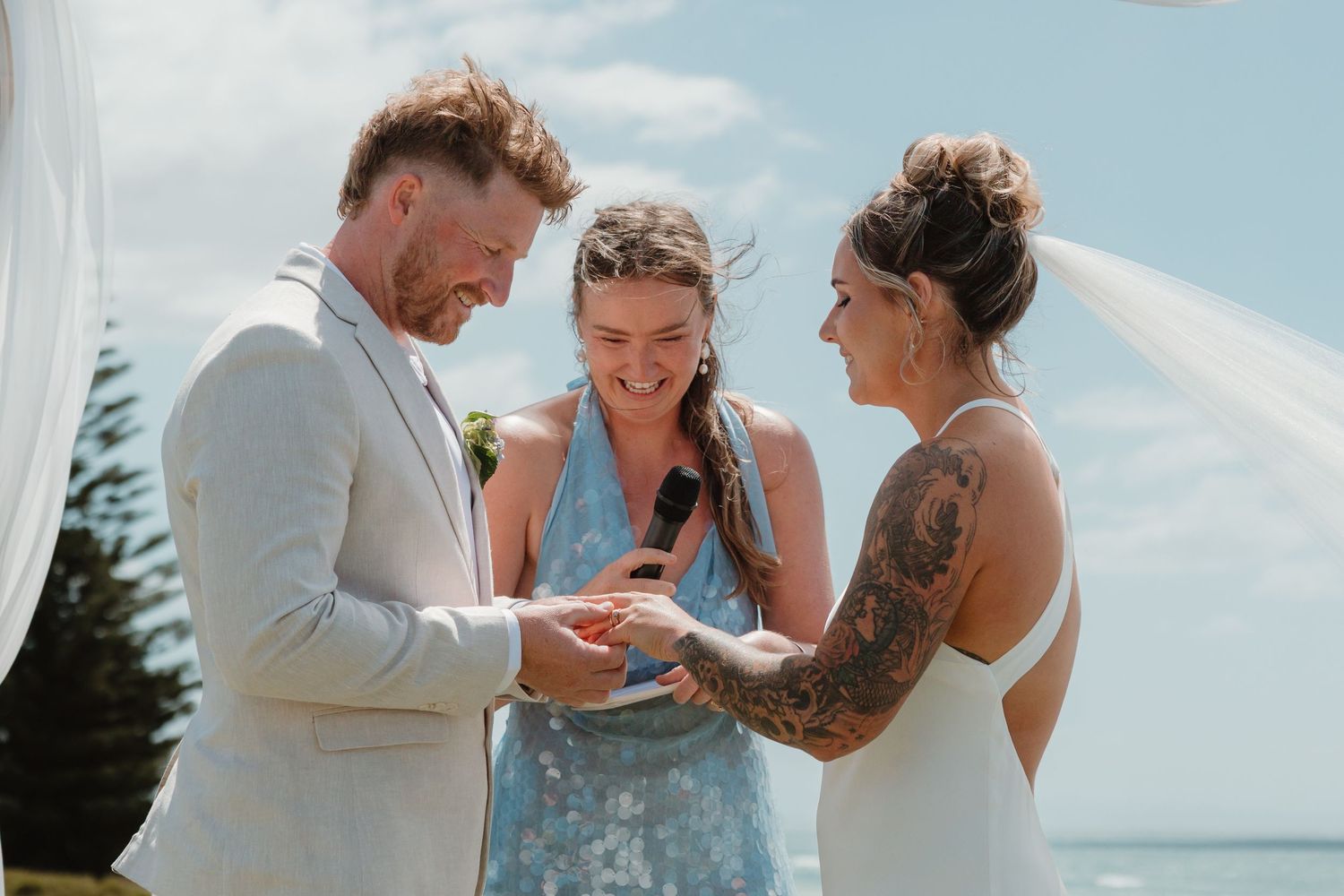 Groom placing wedding ring on bride's finger during outdoor beach ceremony with officiant in blue dress.