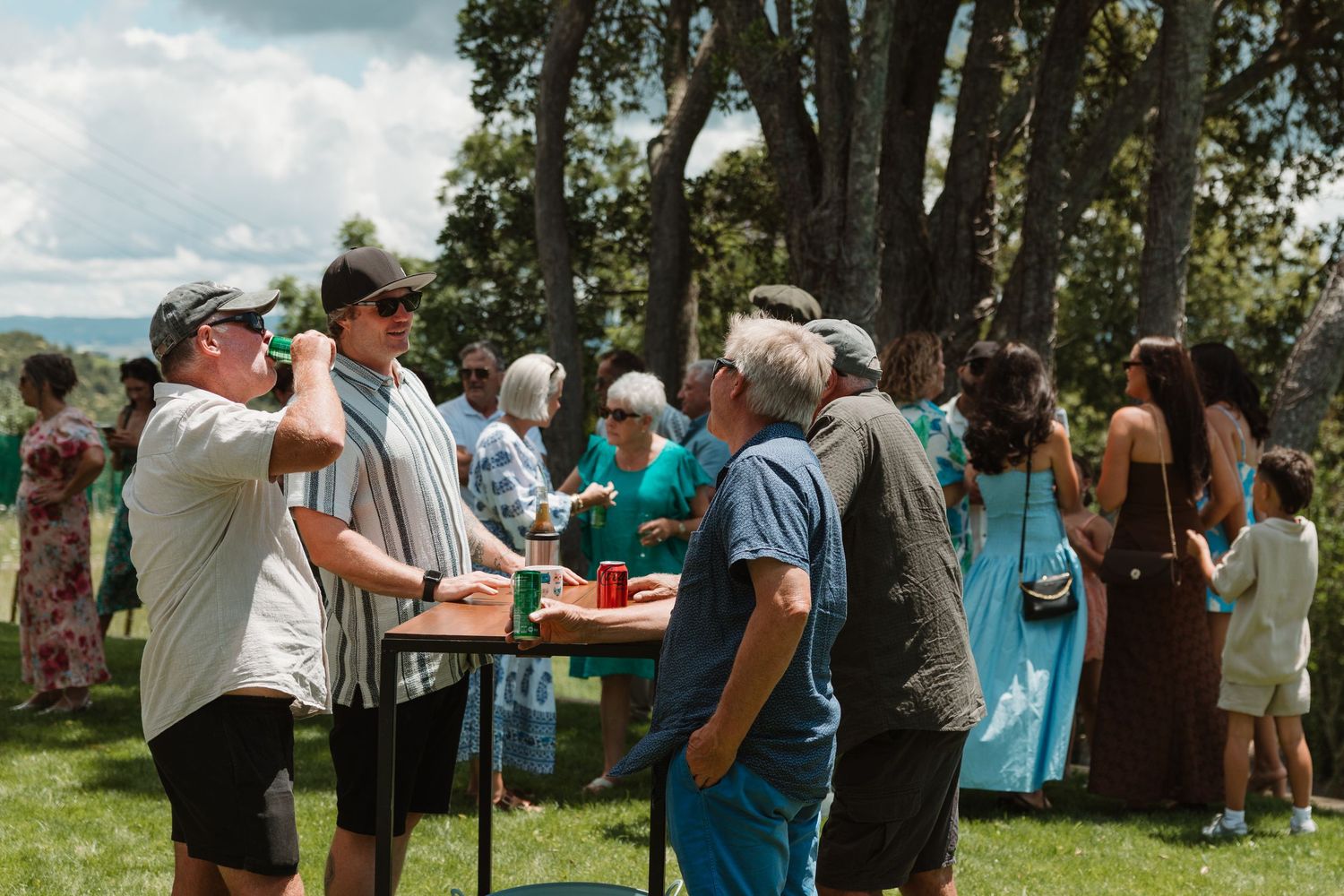 Guests mingle outdoors at a summer gathering on green grass with trees and cloudy skies in the background.