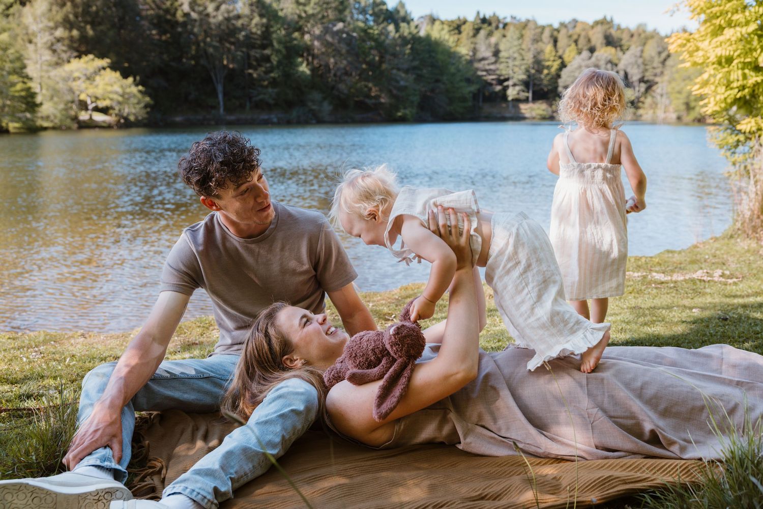 Family relaxing by a lake on a sunny day, with parents lying on a blanket and children playing nearby.