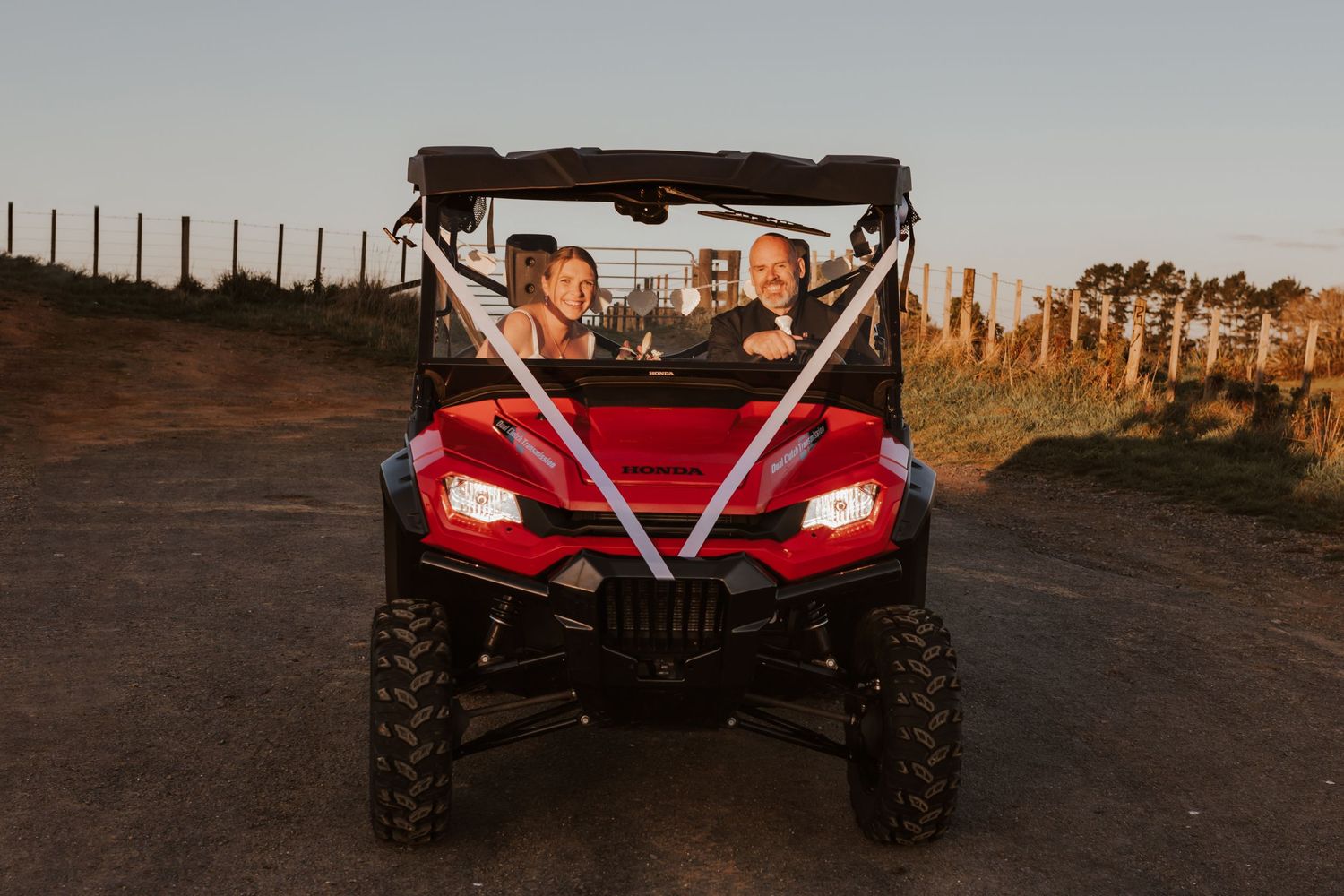 Red utility vehicle with decorative ribbon driving on dirt path at sunset.