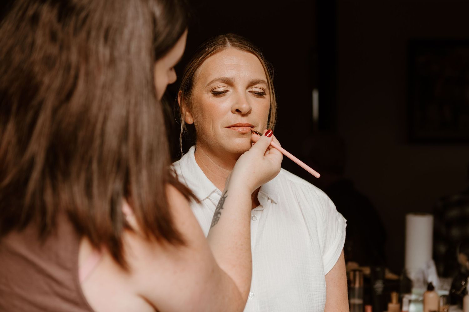 A makeup artist applies lipstick on someone wearing white during a beauty preparation session in warm lighting.